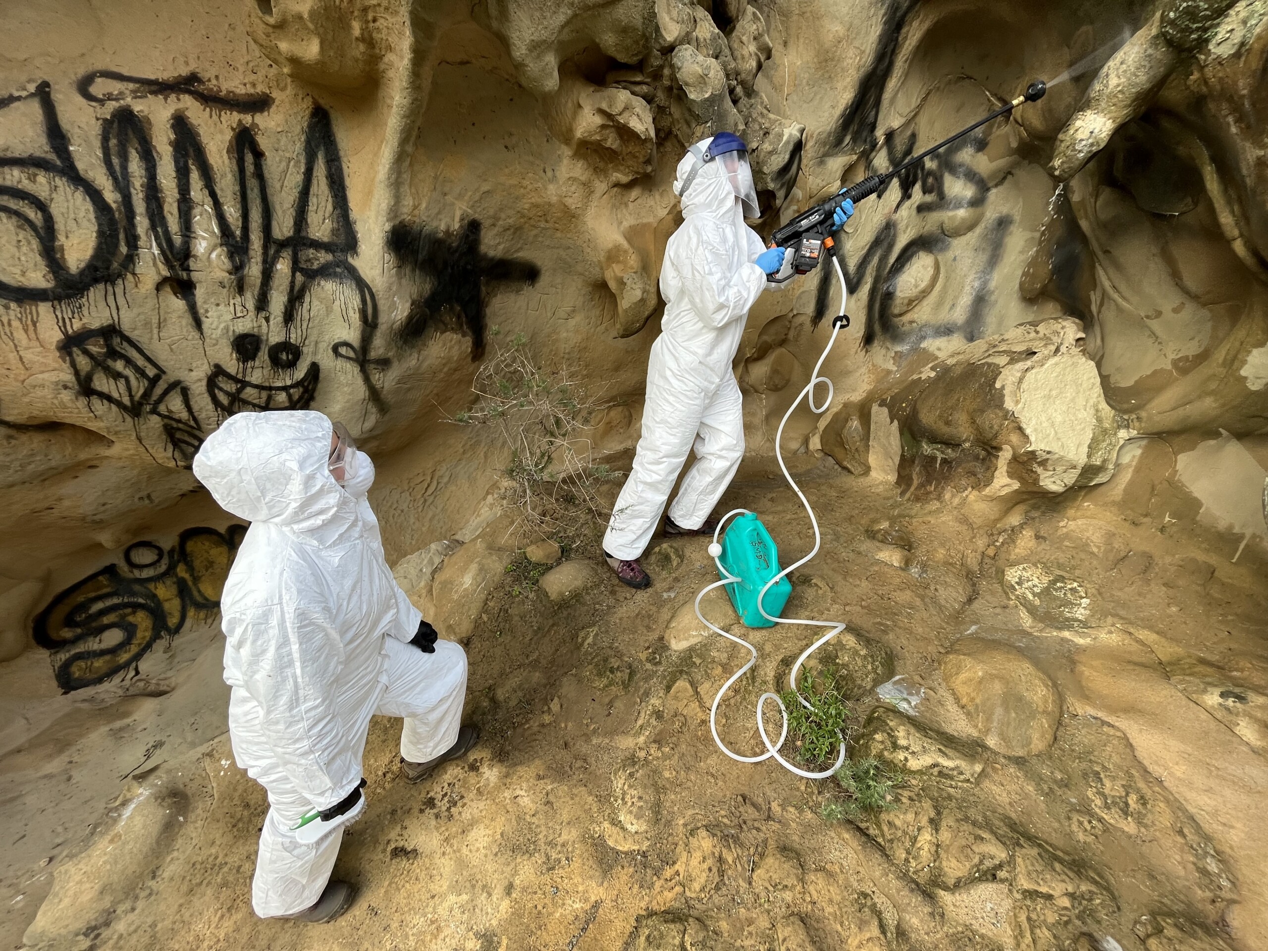 Two people using a power washer to clean graffiti off rocks