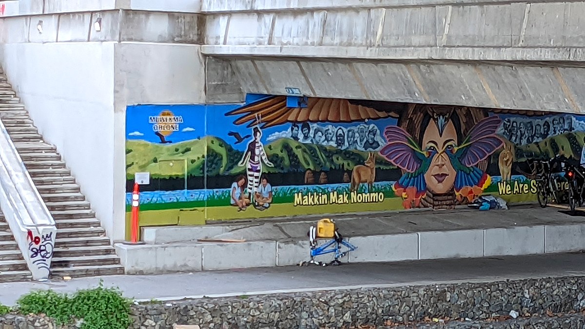 a mural depicting the face of Muwekma Ohlone Chairwoman Charlene Nijmeh adorned in a warrior eagle headpiece and a hummingbird warrior face mask paying tribute to the tribe's creation story