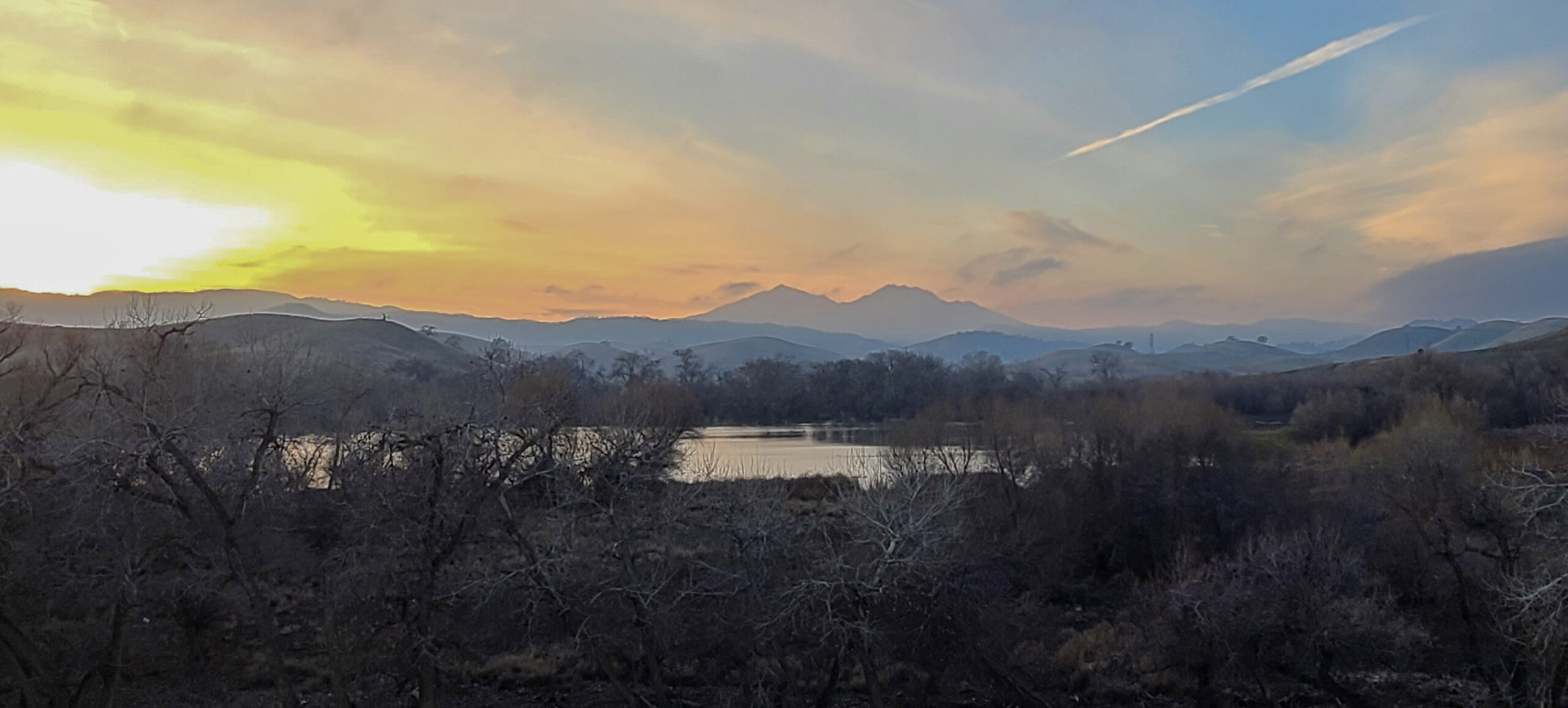 Marsh Creek Reservoir in the foreground surrounded by trees with a sunset showing off the dimension of Diablo foothills in a hazy evening.