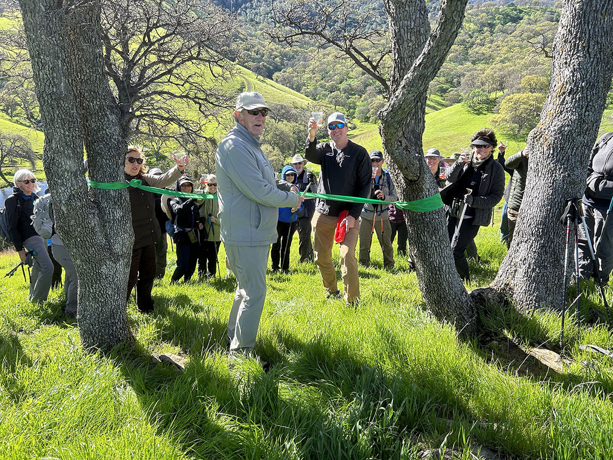 A smiling group of hikers toast a ribbon-cutting on a sunny green hillside